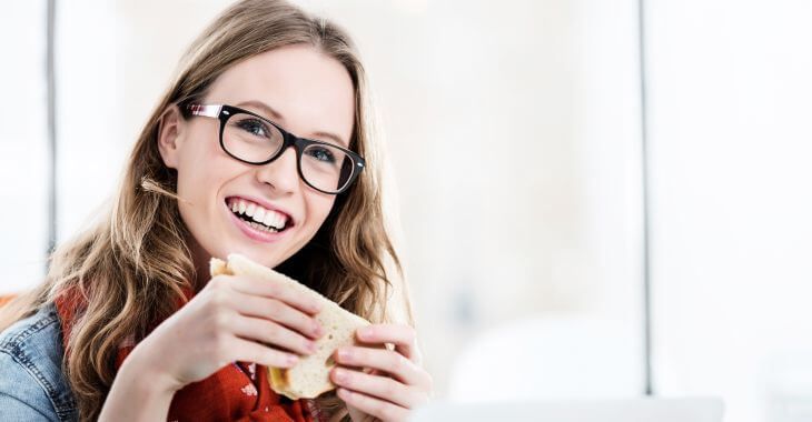 A cheerful young woman with glasses eating a sandwich.