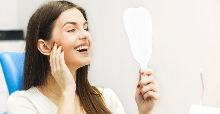 Satisfied woman after dental restoration with mini dental implants looking at her teeth in a mirror.