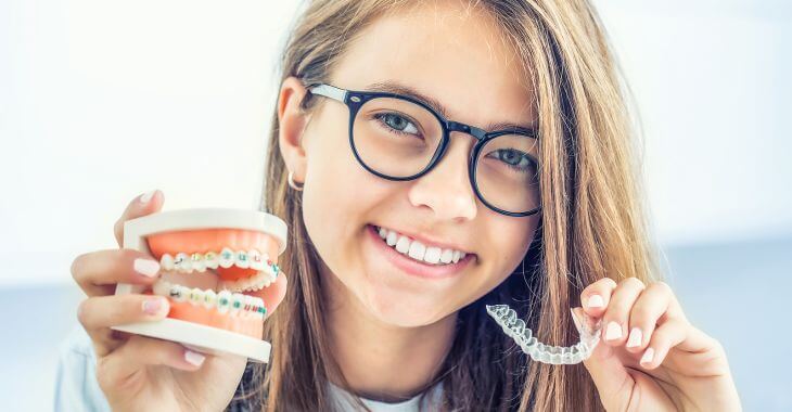 A teenage girl holding a dental model with braces in one hand and a retainer in the second hand.