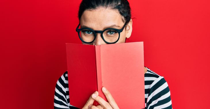 Smiling young woman hiding her mouth behind an open book because of teeth stains.