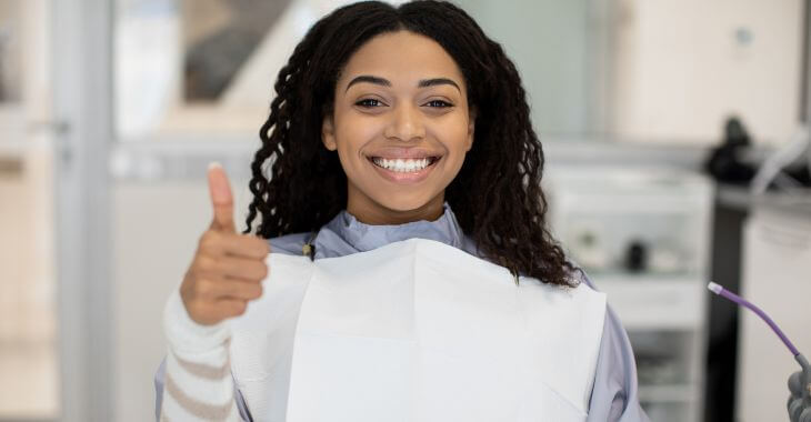 Happy Afro-American woman with perfect smile sitting in a dental chair and showing her thumb up.