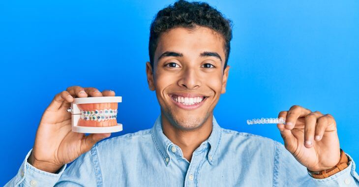 A teen Afro-American boy with perfect smile holding a dental model with braces in one hand and a retainer aligner in the second hand. 