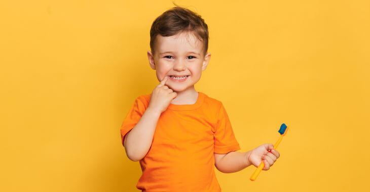 Happy baby boy with a toothbrush in one hand pointing at his teeth.