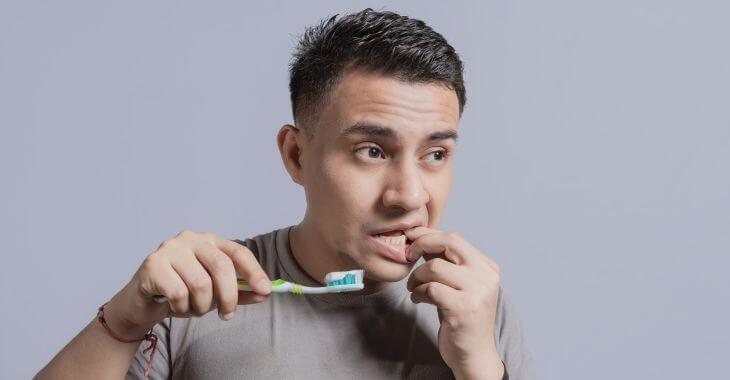 A concerned man with holding a toothbrush and touching his gums with the other hand.