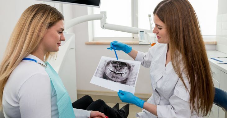  dentist with teeth imaging explaining treatment to a woman patient sitting in a dental chair.