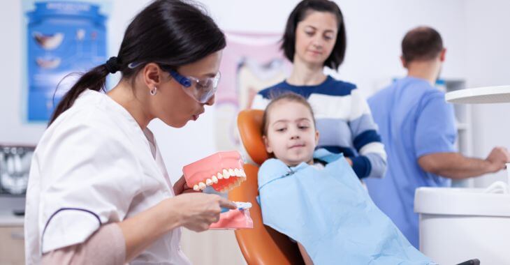 Dentist with a dental model explaining the issue to a mother and her little daughter sitting in a dental chair.