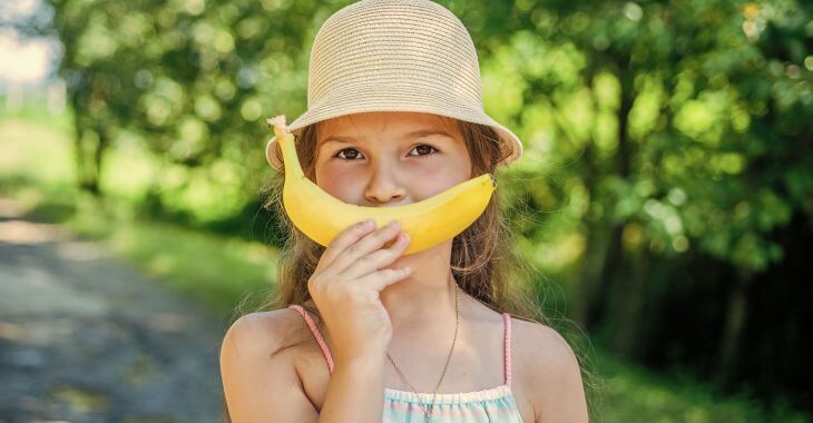 A little girl holding a banana in front of her face and covering her mouth with it.