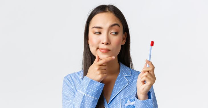 A thoughtful young woman wearing pyjamas looking at a toothbrush not sure if to use it.