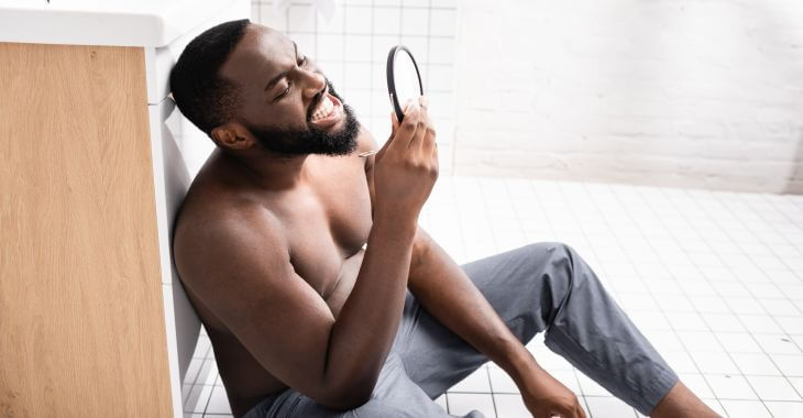 A concerned Afro-American man sitting in the floor n the bathroom and looking at his teeth and gum in a mirror. 