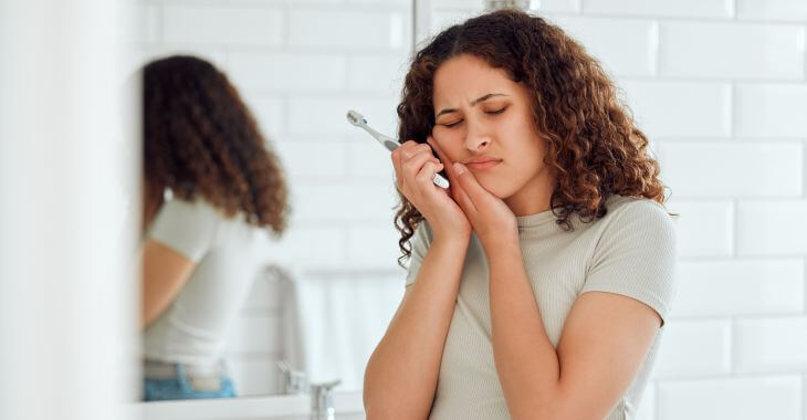 Young woman with a severe toothache standing in front of the bathroom mirror holding a toothbrush in her hand and pressing her jaw an cheek with other hand