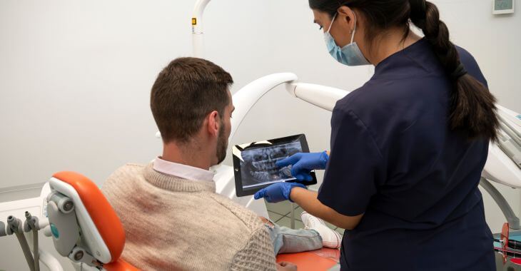 A dentist holding a tablet with digital dental imaging and explaining the issue to a male patient sitting in a dental chair