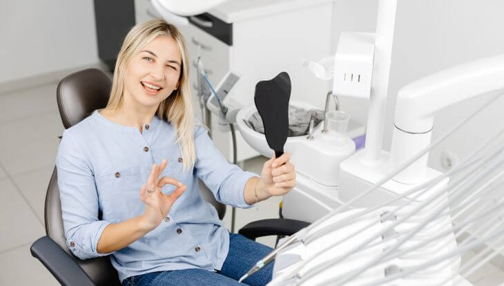 A woman happy with dental restrative treatment results holiding a mirror and showing ok with her fingers while sitting in a dental chair after the procedure
