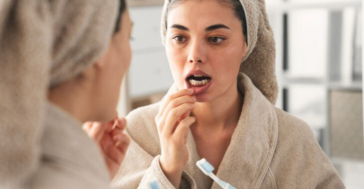 A worried woman wit a toothbrush and towel over her hair looking closely at her gums in a bathroom mirror