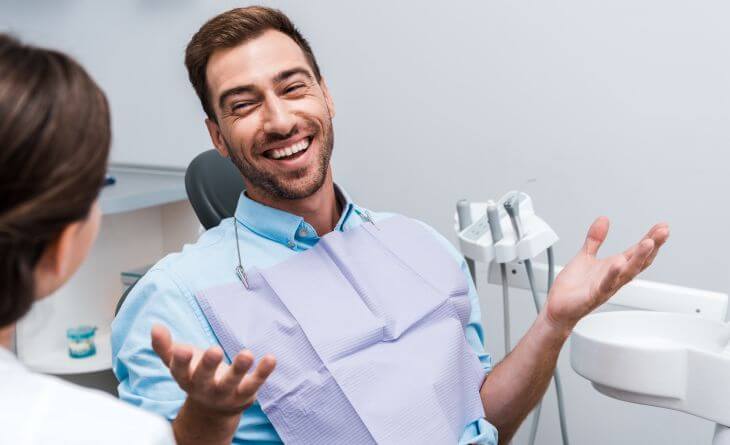 A happily smiling male patient sitting in a dental chair and talking to a dentist