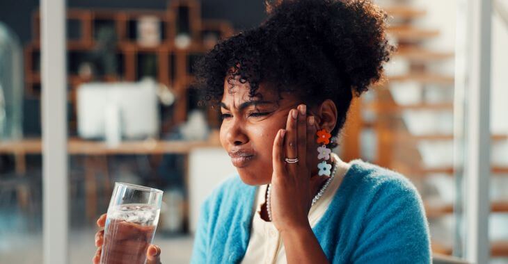 An Afro-American woman with glass of water feeling pain due to teeth sensitivity after taking a sip of water